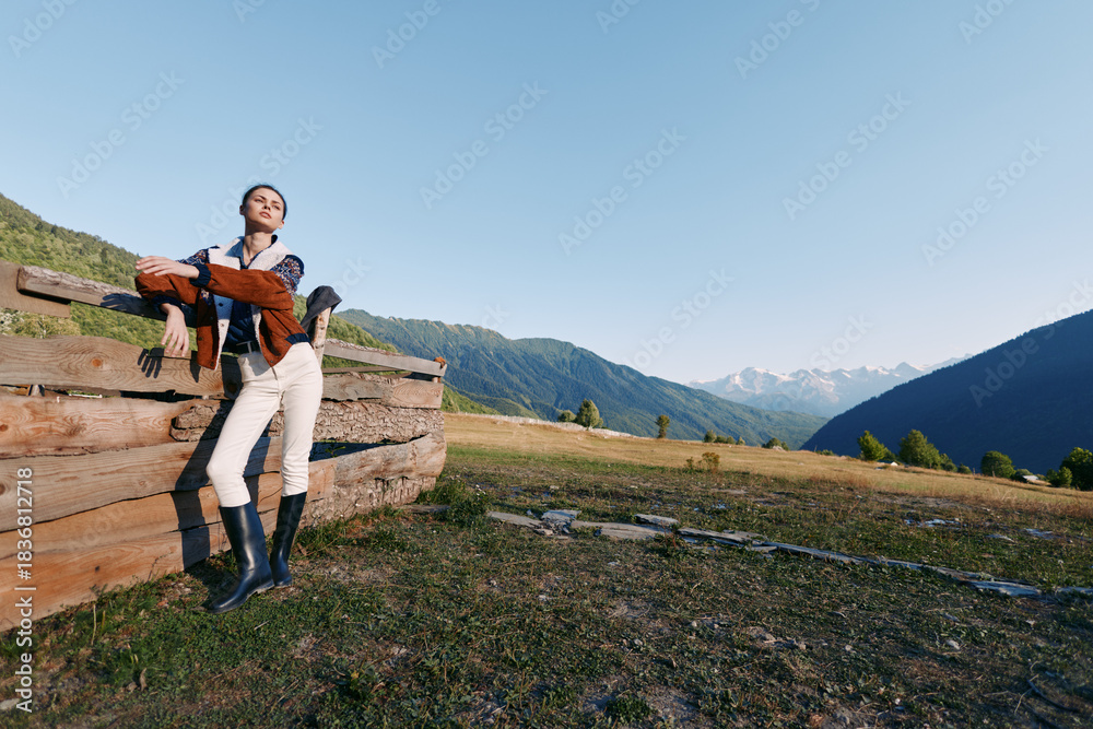 Obraz premium Woman leaning on wooden fence in sunny mountain meadow, wearing riding boots and holding jacket, relaxed countryside portrait with expansive alpine view and clear blue sky.