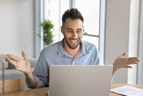 Happy Hispanic man sits at table looking at laptop screen and gesturing during videoconference event, reacting enthusiastically on good news in e-mail or notifice, celebrating results, feels excited
