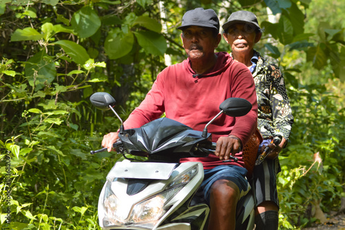 A man in a red shirt and a woman in a patterned black and gold top pause on a white scooter along a rural path surrounded by dense tropical foliage, looking directly at the camera.