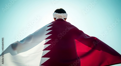 Person with traditional headwear holding a large Qatar flag against a bright sky, symbolizing national pride and identity.