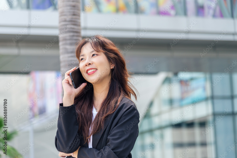 Fototapeta premium Young Asian business woman, busy with work on street, wearing black suit talking on phone with happy smile, enjoying modern urban cityscape, professional working outdoors
