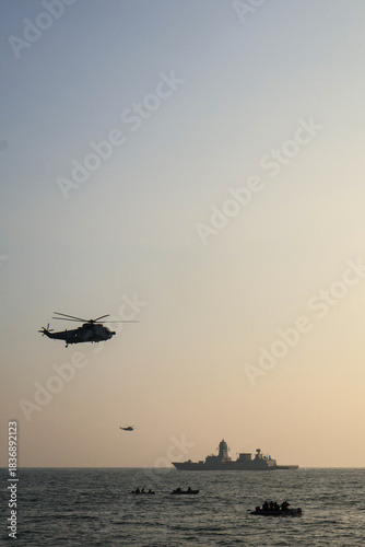 Bild auf Leinwand Trivandrum India December 3 2025 : Indian Navy helicopter and warship operate together at sunset off Shangumugham Beach with small boats in foreground during Navy Day demo
