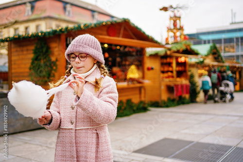 Little cute kid girl eats sweet cotton candy on Christmas market. Happy child on traditional Xmas fair in Germany.
