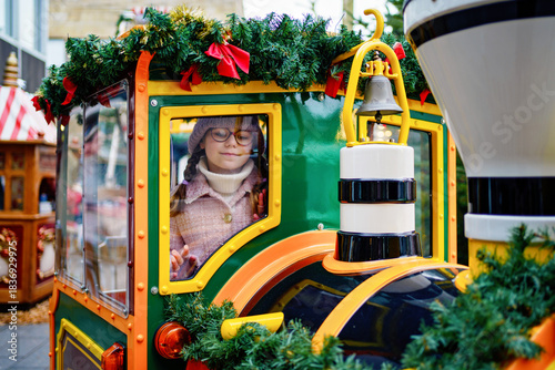 Little school girl on a carousel train at Christmas funfair or market, outdoors. Happy child having fun. Traditional xmas market in Germany, Europe. Holiday, children, lifestyle concept