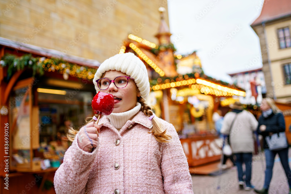 Obraz premium Little cute kid girl eat sweet sugar covered red candy apple on Christmas market. Happy child on traditional Xmas fair in Germany.