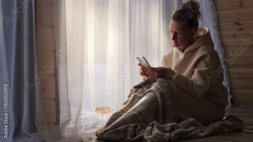 Woman wrapped in blanket sits by window with glass of wine scrolling through her phone Calm light blends silence with glow of screen Phone combines calm symbol of mindful telephonic contemplation