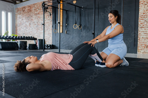 Two women doing situps on black mats in CrossFit gym while trainer stabilizing ankles by dumbbells