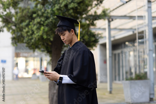 Asian male graduate standing in courtyard wearing gown and mortarboard looking at smartphone