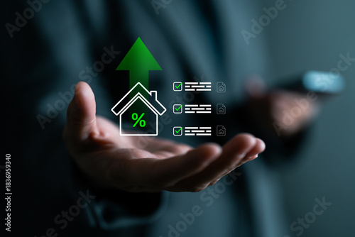 Close-up of hand with digital overlay of house, percentage symbol, checklist and upward arrow. Represents rising property value, real estate investment, mortgage rates or home loans.