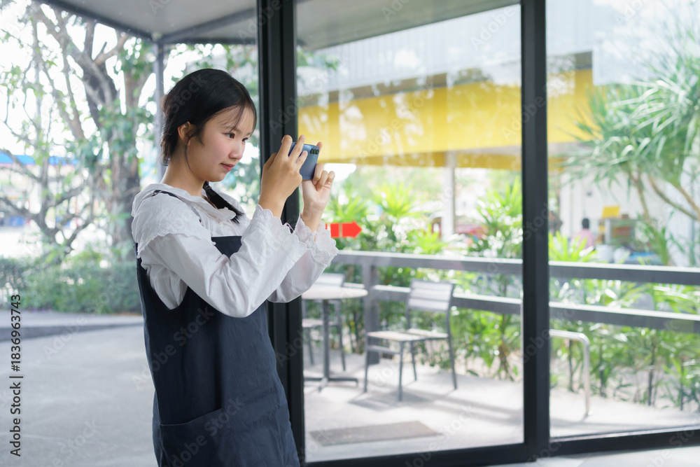 Fototapeta premium Teenage girl standing and taking a photo with her smartphone inside a café, expressing creativity, focus, and modern digital lifestyle in a bright natural environment