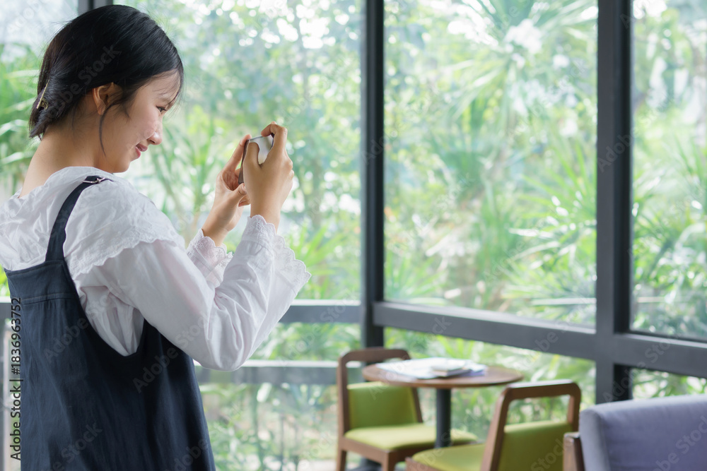 Fototapeta premium Teenage girl standing and taking a photo with her smartphone inside a café, expressing creativity, focus, and modern digital lifestyle in a bright natural environment