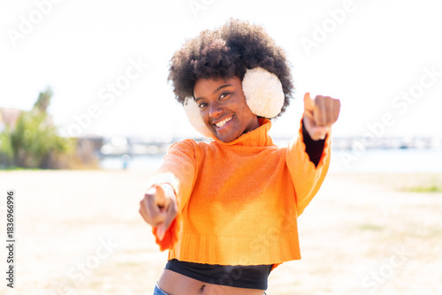 African American girl wearing winter muffs at outdoors points finger at you while smiling