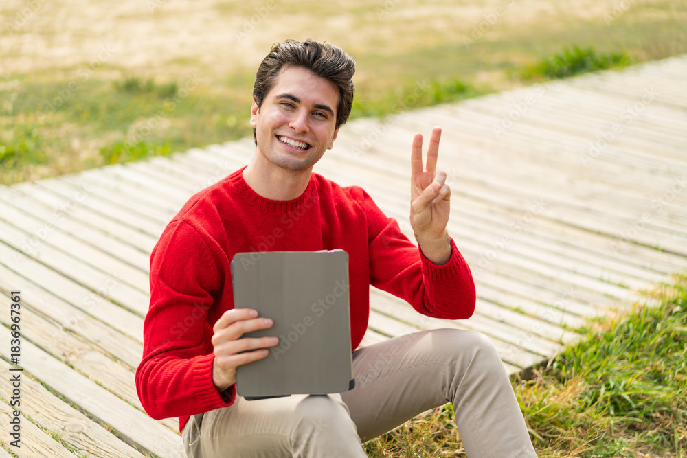 Fototapeta premium Young handsome man holding a tablet at outdoors smiling and showing victory sign