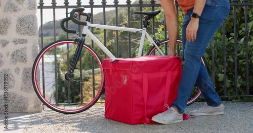 courier approaching bicycle and bag on walkway, lifting bag onto back and pulling bike for delivery