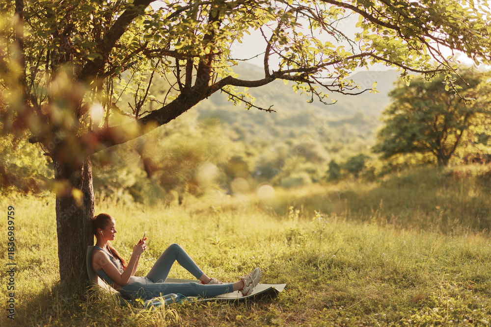 Obraz premium Young woman relaxing under a tree in a sunlit meadow, enjoying her phone amidst the serene natural landscape, representing tranquility and leisure in nature