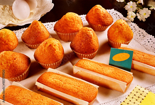 A rustic wooden table holds a vibrant spread of pastries. Freshly baked almond cupcakes with golden peaks sit next to a flaky, buttery croissant, offering a delightful contrast of textures and a warm,