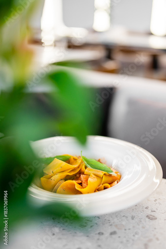 A close-up, selective focus shot of a gourmet pappardelle pasta dish in a white bowl, garnished with fresh basil leaves and served in a modern restaurant.