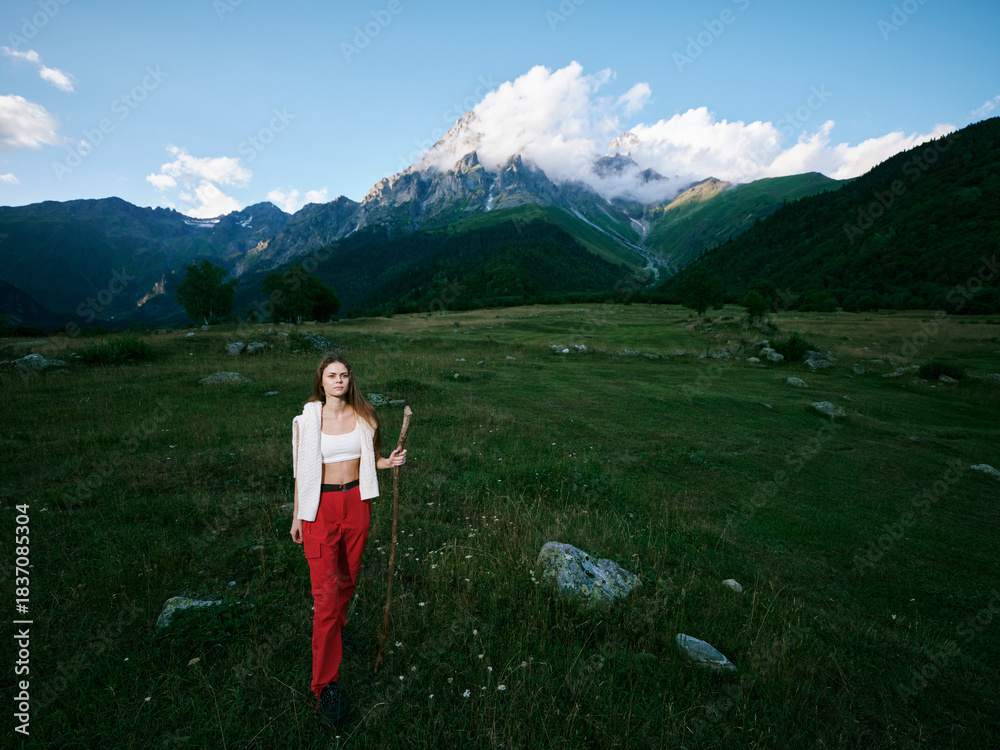 Obraz premium Woman standing in a grassy meadow with distant mountains and blue sky, wearing a white cardigan and red pants, holding a wooden staff in a scenic outdoor landscape