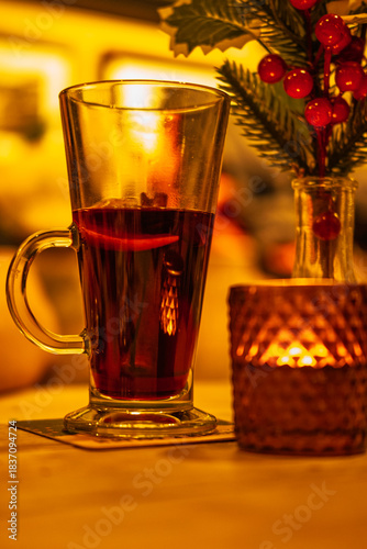 A glass of mulled wine sits next to a lit candle and a sprig of holly, creating a cozy and festive atmosphere.