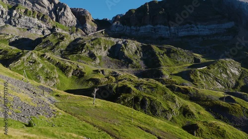 Aerial footage moving over a stunning mountain pass in the pyrenees, showing lush green slopes, rocky peaks, and a clear blue sky on a sunny day