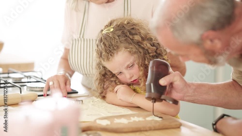 Cute curly-haired girl watching her grandfather pour chocolate frosting on homemade cookies. Family spending quality time together baking