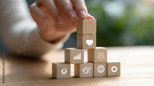a person stacking wooden cubes into a pyramid shape, each cube has an icon: heart, gear, graph, globe, on a light wood table, natural daylight