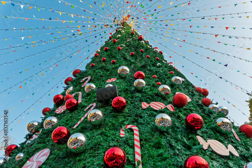 Giant, brightly decorated outdoor Christmas tree, viewed from below, with red and silver ornaments and festive pennant strings against a blue sky.