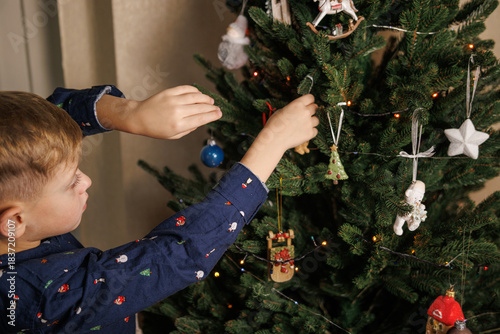 The process of decorating a Christmas tree - a child in a shirt holds ornaments and hangs them on the tree.

