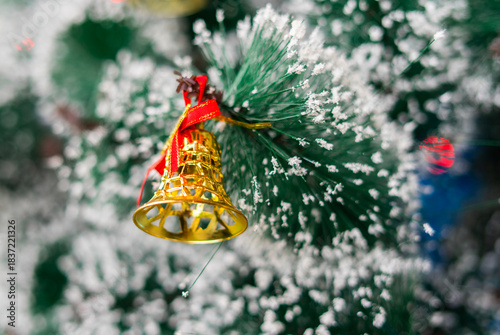 A tiny golden bell with a red ribbon hangs on a snow-dusted Christmas tree branch. A classic and festive holiday decoration that adds sparkle to the season.