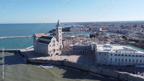 A panoramic coastal view of Trani shows the famous seaside cathedral overlooking turquoise waters, a long breakwater with lighthouses, and the historic old town bathed in bright Mediterranean sunlight