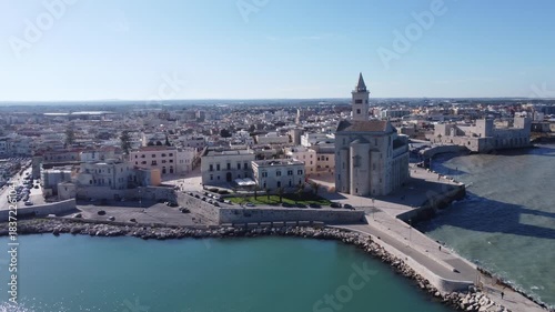A panoramic coastal view of Trani shows the famous seaside cathedral overlooking turquoise waters, a long breakwater with lighthouses, and the historic old town bathed in bright Mediterranean sunlight