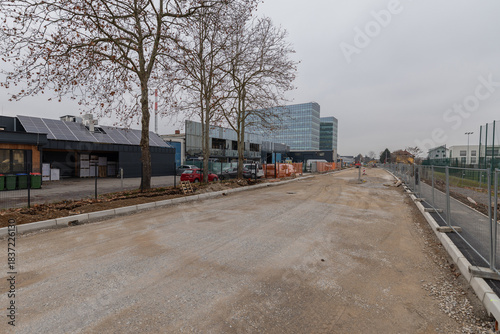 Active construction site on Magistrova street, Ljubljana, featuring an orange excavator, deep trench with concrete walls, workers laying utility pipes, and residential buildings under a cloudy sky.