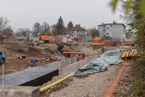 Active construction site on Milcinskega street, Ljubljana, featuring an orange excavator, deep trench with concrete walls, workers laying utility pipes, and residential buildings under a cloudy sky.