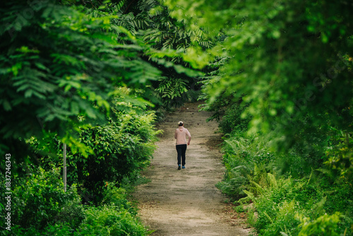 A lone figure runs down a dirt path through a lush, green forest. The path leads into the distance, framed by dense foliage. It evokes a sense of solitude and journey.