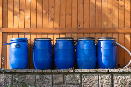 Fotografie Five blue plastic barrels with black lids are neatly lined up on a stone platform beside a wooden fence