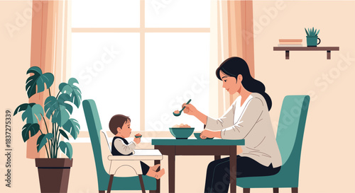 Caring mother sits at a dining table and patiently feeds her young child who is sitting in a high chair at home.