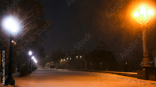 Fabulous view of the snow-covered city park at night, with trees in the snow, cozy warm light of lanterns.
