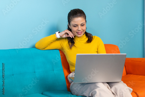 Woman wearing yellow turtleneck sitting on two-tone sofa in office using laptop and smartwatch