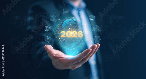 A businessman holding a glowing holographic globe with 2026 and global economy icons, symbolizing worldwide market trends, currency exchange, trade, logistics, and economic growth insights for 2026.