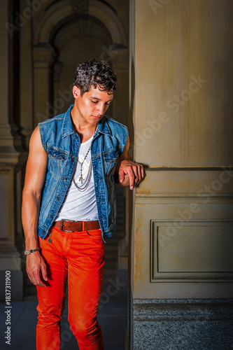 Stylish Man in Denim Vest. A young man leans against a wall in a thoughtful pose, wearing a denim vest over a white shirt and bright orange pants. The setting is an elegant architectural space.