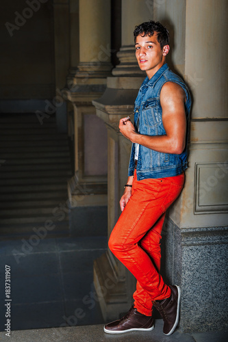 Casual Fashion in Urban Setting. A young man stands confidently against a stone ledge. He wears a denim vest and bright red pants, creating a bold contrast against the muted architectural background.