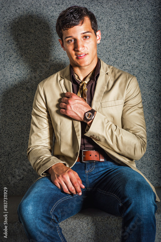 Stylish Young Man in Casual Attire. A young man sits confidently on a concrete bench. He wears a beige blazer, brown shirt, and blue jeans, accessorized with a watch.