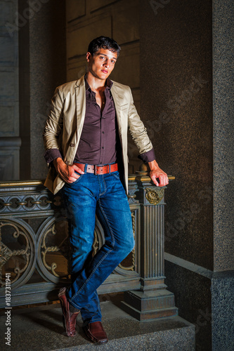 Stylish Man Leaning Against Railing. A man in a beige jacket and jeans leans casually against an ornate railing. The setting is nighttime, with shadows and warm lighting creating a dramatic effect.