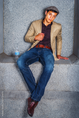 Man Sitting in Casual Attire. A young man sits casually against a stone backdrop, wearing a beige jacket and blue jeans. A bottle of water sits nearby, adding to the relaxed vibe.