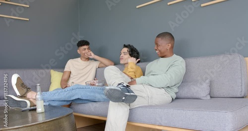 Diverse male friends lounging on couch, eating chips, laughing after inspecting friend picking nose
