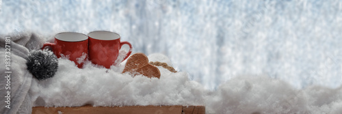 two red mugs with wooden  heart shaped arranged on the snow with blanket on a plank and in front of frozen glass of a window  with copy space on the right