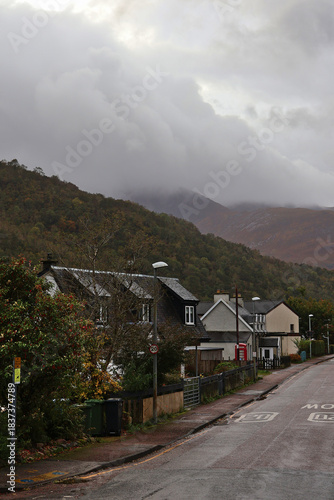 Gleann Comhann village high street in Glencoe, Scottish Highlands; quaint village scene with mountains rising through fog in distance and red British postbox in midground