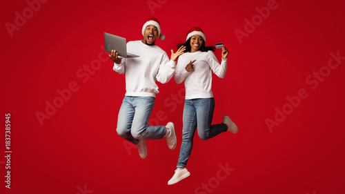 Two people are joyfully jumping in front of a bright red background. They are wearing festive hats and holding a laptop in one hand and a credit card in the other hand, showcasing holiday spirit.