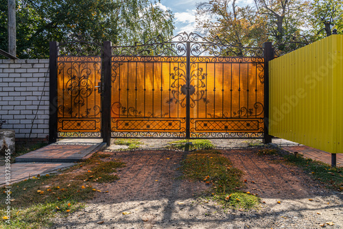 Closed openwork metal gate with orange transparent plastic in the daytime close-up on a private land plot interior view with shadow.