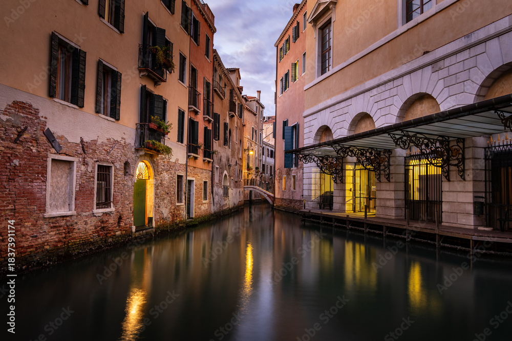 Fototapeta premium Evening in Venice - calm surface of a small canal between houses with reflection of lights on the surface